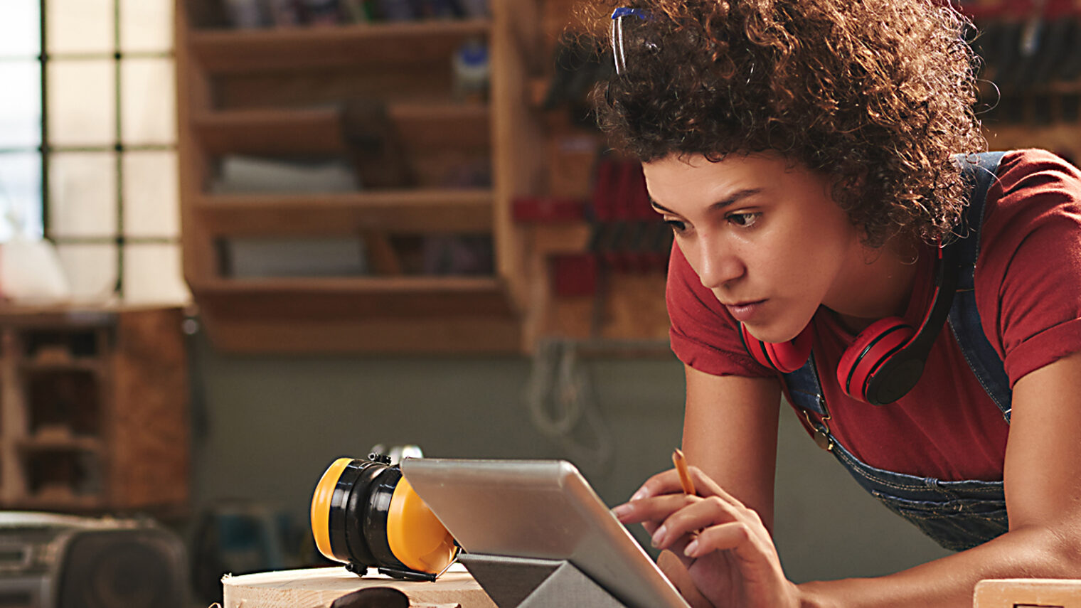 Young concentrated woman with curly hair reading instructions on digital tablet before working with wood Schlagwort(e): woman, female, young, attractive, curly, concentrated, digital, tablet, technology, internet, leaning, table, looking, reading, wifi, carpentry, joinery, timber, wood, wooden, plank, tool, skill, manual, equipment, professional, craft, craftsman, handyman, occupation, working, woodworking, workshop