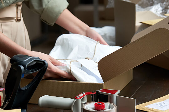 Frau verpackt zwischen vielen Paketen und Umschlägen ein papiergebundenes Päckchen in ein Karton.