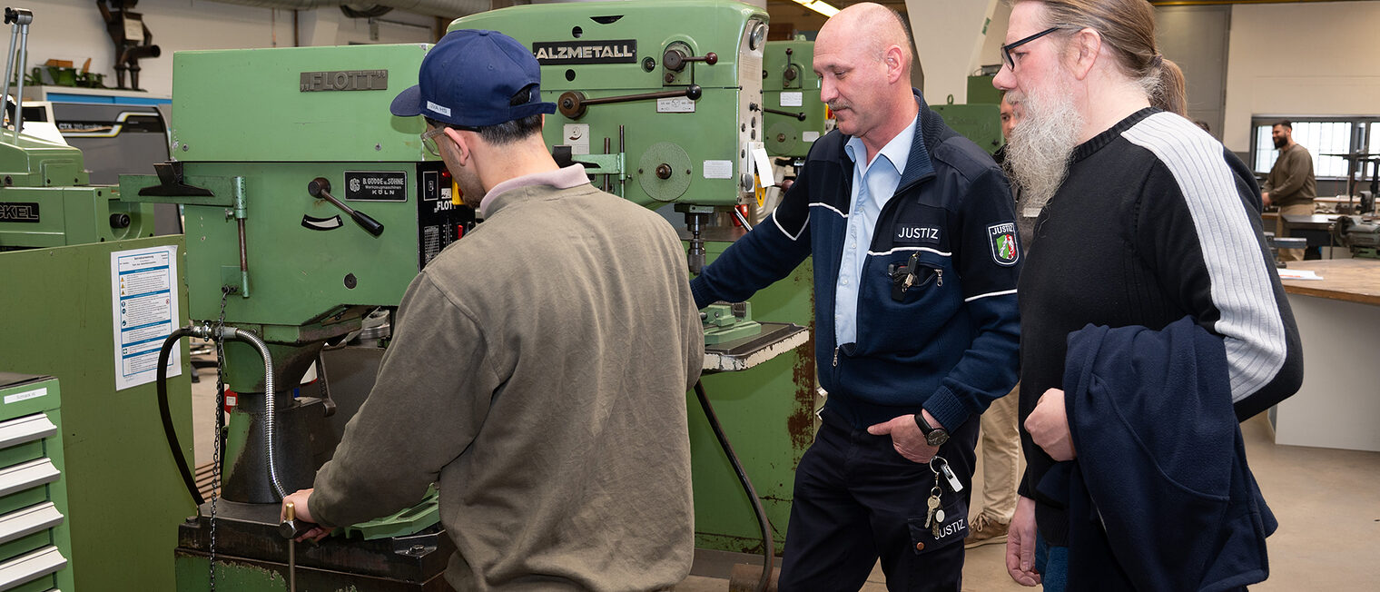 Der Häftlinge steht an einer Bohrmaschine. Neben ihm stehen ein fachlicher und ein sozialpädagogischer Betreuer.