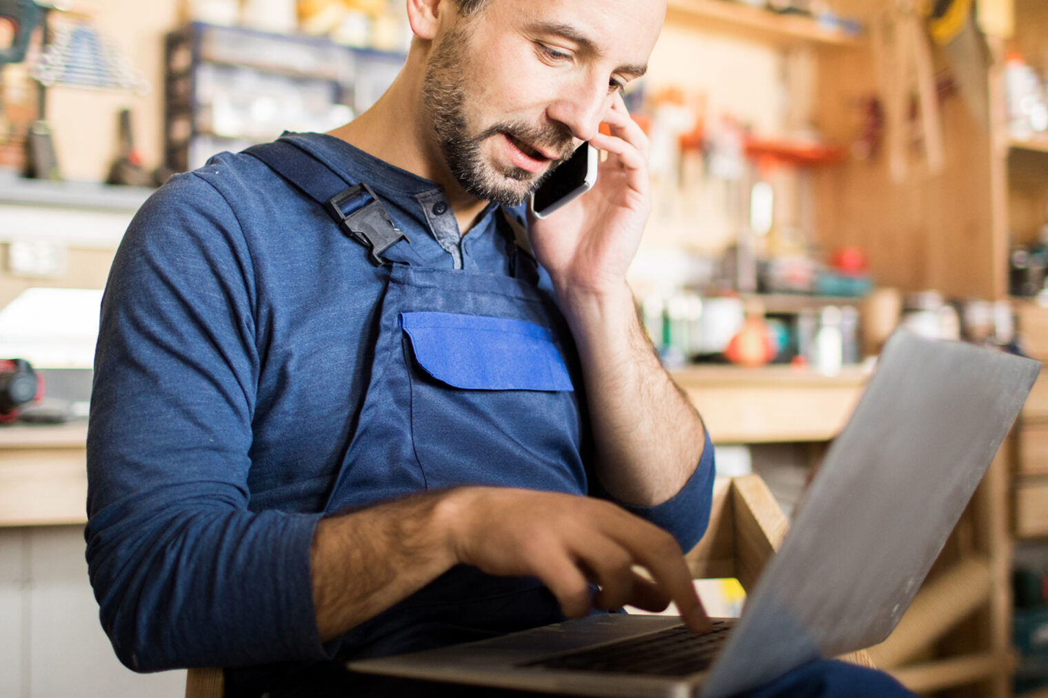 Porträt eines Mitarbeiters, der im Werkstattbereich einen Laptop benutzt und über das Smartphone spricht.