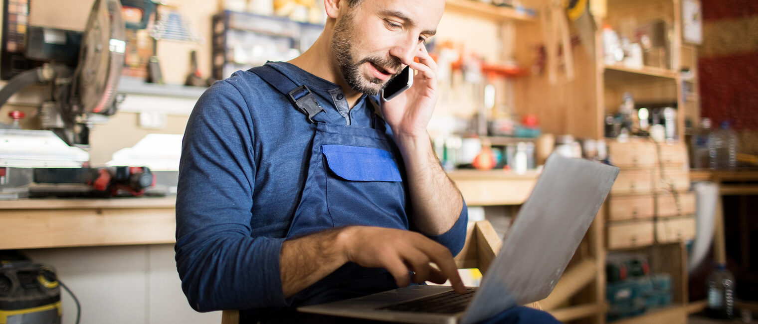 Porträt eines Mitarbeiters, der im Werkstattbereich einen Laptop benutzt und über das Smartphone spricht.
