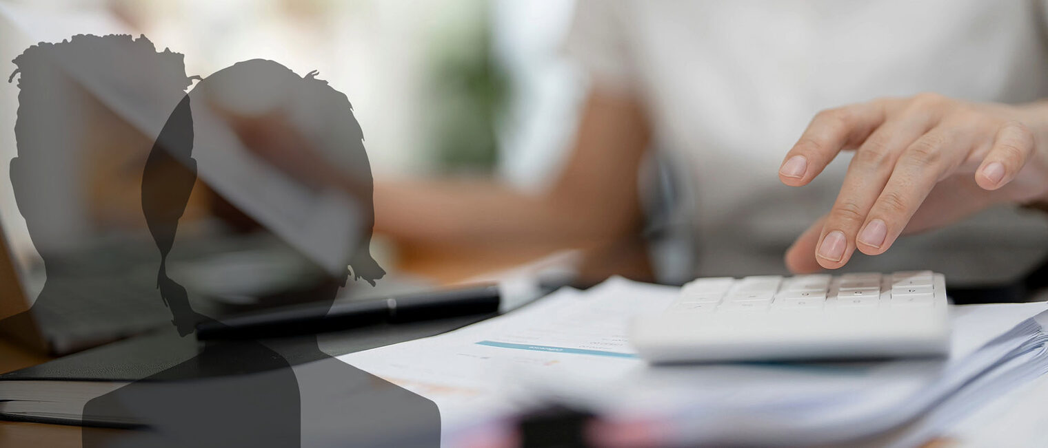 Bild zeigt eine Frau, die im Büro arbeitet und Unterlagen, einen Rechner und ein Laptop bedient.